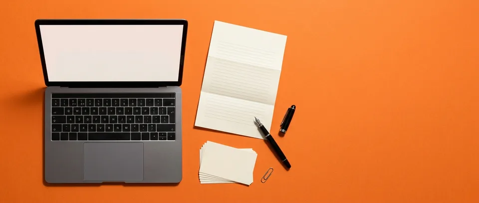 An open laptop, a folded letter, a fountain pen and blank index cards on an orange desk